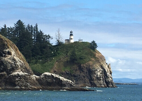 Lighthouse perched on green bush covered rock cliff overlooking the ocean, blue sky above with layered white clouds.