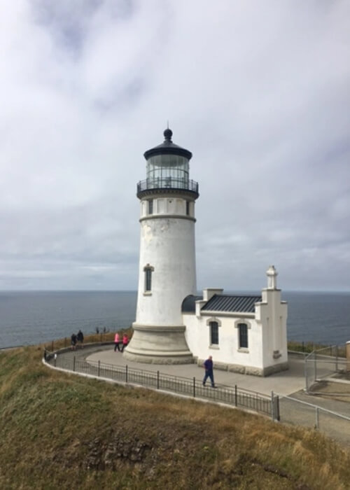 alt="Lighthouse with small side building on side of hill overlooking ocean, grey skies above. A few people walking around base of lighthouse."