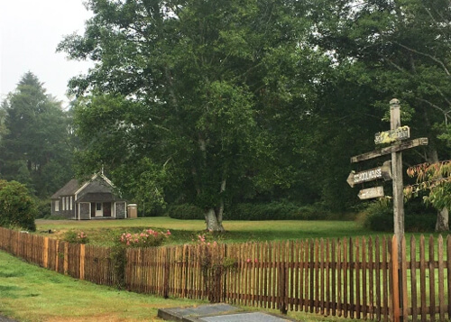 Picket fence covering the length of the bottom of picture, leading to early 1900's school house at left. Wooden sign pointing to school house at right.  School grounds covered in grass and trees, grey sky.