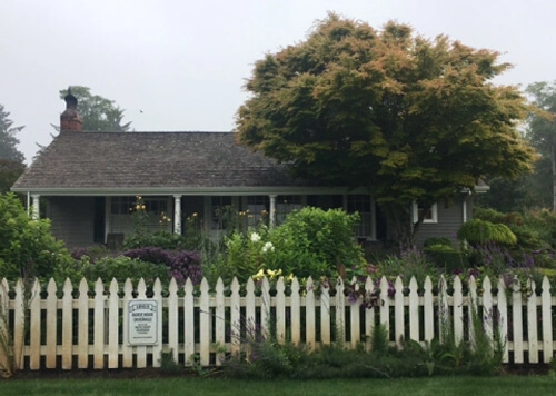 White picket fence in front of very plain single story house with trees and bushes in front yard, sign on fence saying "oldest house in Oysterville - 1863."