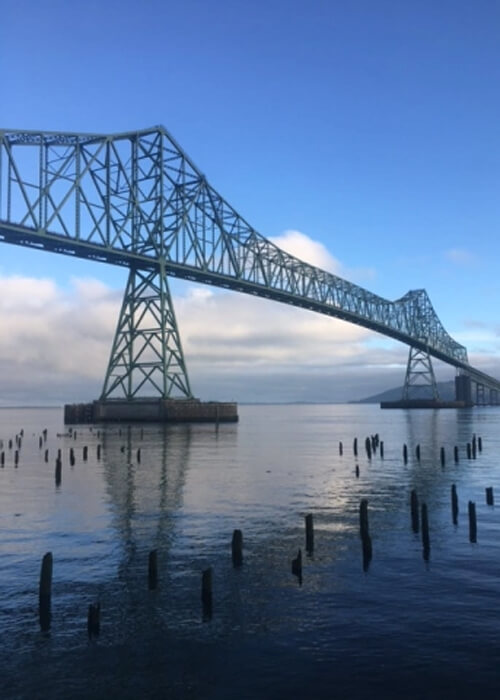 Large green steel bridge crossing body of water covering the length of photo.  White clouds and blue sky reflecting in still water below.