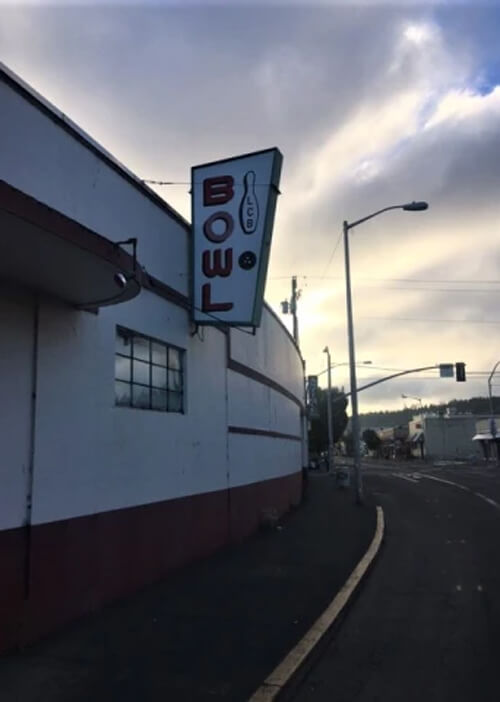 Sign reading "Bowl" with bowling pin on its perched on side of white painted round cement building.  Thick clouds above with a little sun coming through above the building.