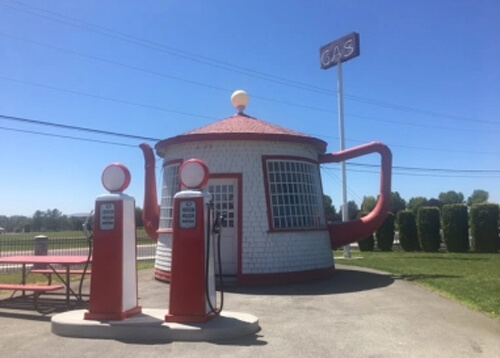 Life-sized teapot with red lid and handle, body painted white.  Two 1930's style gas pump replicas in front of it, blue sky in background.