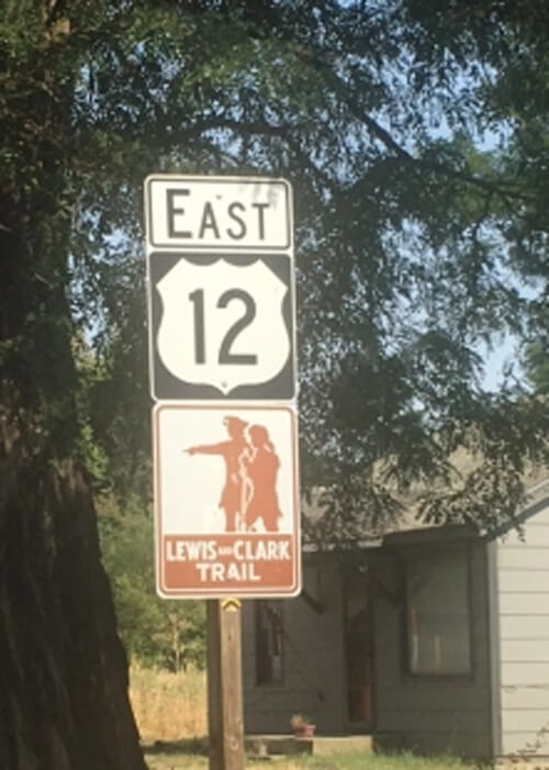 Highway sign reading "East, 12, Lewis and Clark Trail" with a tree and house behind it.