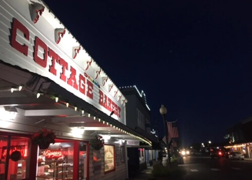 Night photo with bakery at left, painted white with big red lettering reading "Cottage Bakery." Dark street at right with a few lights in the distance.