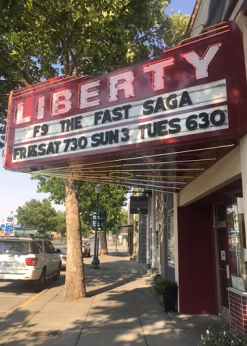 Red neon theatre sign reading "Liberty" with theatre entrance at right and tree overhanging sign above.  Blue sky in background.