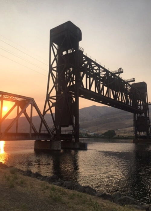 Rusty iron vertical left railroad bridge above river with sun coming through steel frame supports at sundown and mountains in background.