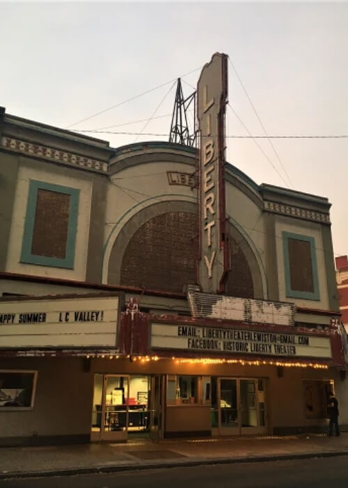 1920's style theatre with vertical sign that reads "Liberty" and marquee displaying movie titles.  Grey sky above.