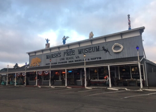 Light blue wooden building with white trim and columns, dark blue sign reading "Marsh's Free Museum." Cloudy sky above and empty parking lot.