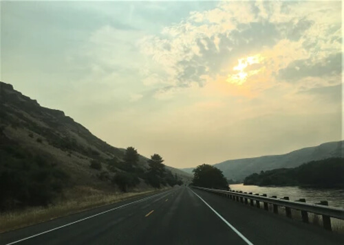 Road disappearing into horizon with river at right and hill at left.  Hazy orange sky.