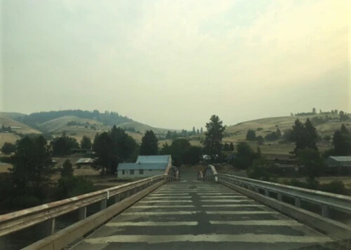 Bridge leading to a few white single story buildings, with low rolling yellow hills and grey blue sky in background.