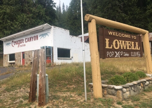 Sign at right reading "Welcome to Lowell" with "population 24" crossed out to read "23."  White brick building at left and dark trees above. 