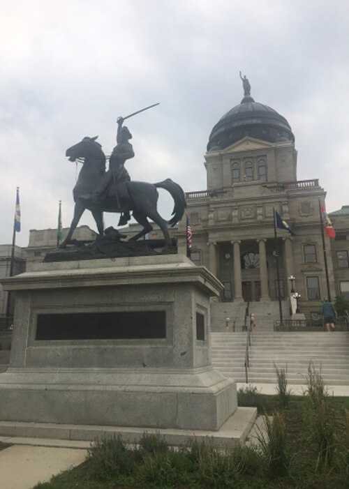 Capitol of Helena cement Greek column style building, with rusty green dome at top and statue of man on horse in front.  Grey sky above. 