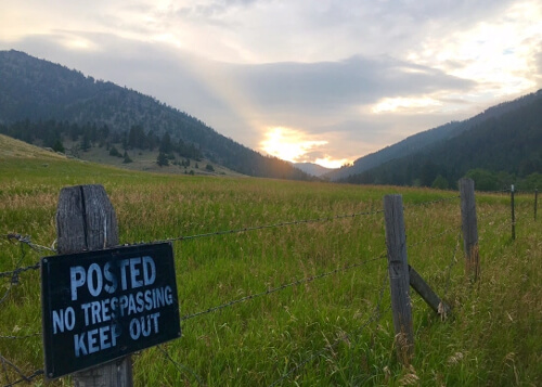 Bright green field with wooden barbwire fence at sunrise with sign reading "no trespassing."  Forested hills and cloudy sky in background.