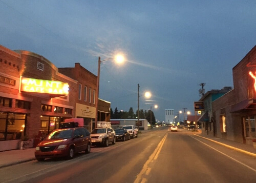 Lighted street just past sundown, with white lit illegible sign at left for restaurant.  Street disappears into the horizon, dark grey sky above.