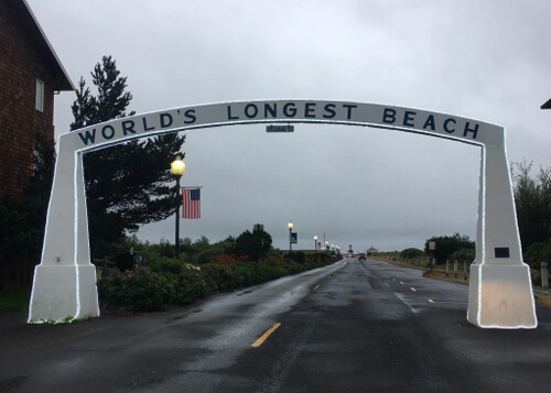 Wet road disappearing into horizon with arch over road reading "World's Longest Beach" and street lights with American flags hanging from them.