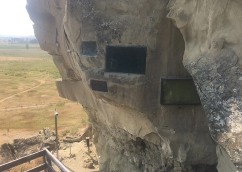 A series of frames protecting rock etchings and signatures around segments of a three-story-high beige-colored rock.  William Clark's signature (of Lewis and Clark) is one of the signatures.