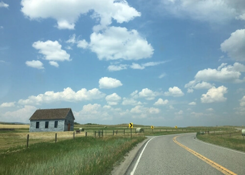 Single story one room house at left in thick green grass and road at right, puffy white clouds in blue sky above.