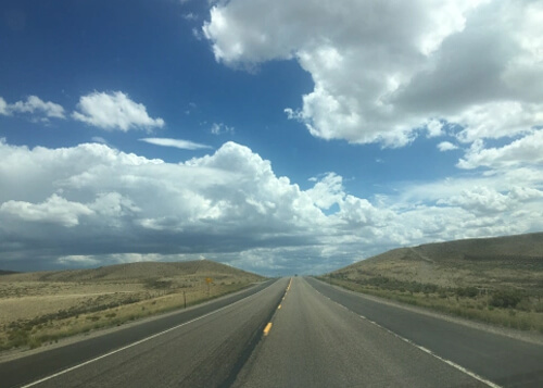 Road disappearing into the horizon, with white pillow shaped clouds and blue sky above.  Grazing grass on both sides of road.