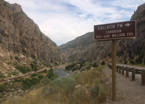 Narrow canyon with light tan rock mountains on both sides and highway sign reading "Gallatin FM Cambrian 500-600 million years" displaying the age of the rock in canyon.