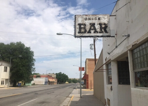 Faded white-painted sign reading "Union Bar" above a white painted building at right, road disappearing into horizon at left, blue sky with scattered clouds above.