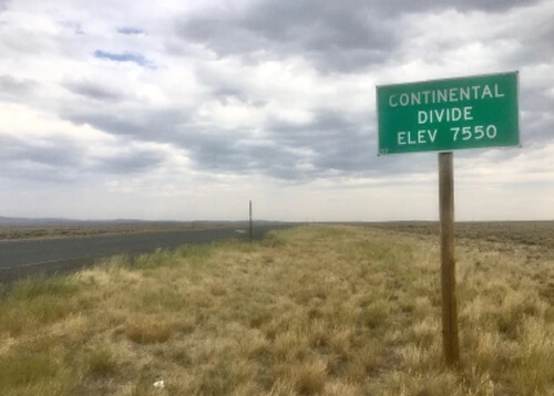 Sign reading "Continental Divide Elevation 7550" at right among a seemingly endless field of yellow grazing grass, cloudy sky above.