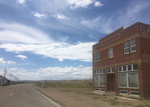 Two story abandoned brick building at right with a series of windows, and street at left disappearing into the horizon. Blue sky with clouds above.