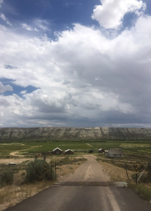 Dirt road leading two a series of red farm houses, with a mountain ridge in distance and huge thick clouds above with some fragments of blue sky.