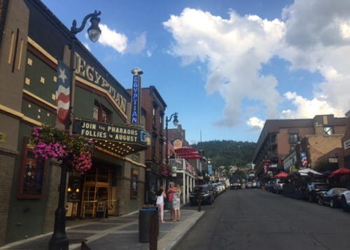 View of a series of downtown single-story buildings on both the left and right of street.  Largest building at left  is painted green and has sign that reads "Egyptian."  Blue sky and some scattered white clouds above.