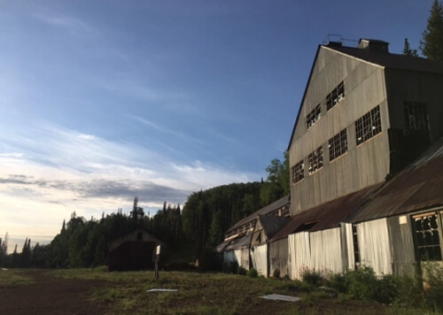 Three-story aluminum mine building at right with a series of broken windows.  Blue sky above and some some green trees behind the mine building.