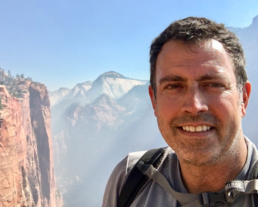 Close-up of Mark Loftin, a smiling middle-aged white male with short dark hair and a view of a mountain landscape behind him.