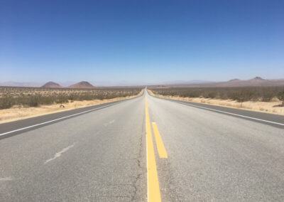 Highway aimed straight ahead through desert terrain, yellow stripes in center of road, blue sky above.