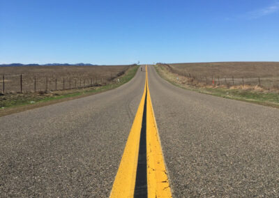 Road aimed straight ahead over a hill, bright yellow stripes in center. Open pasture on both sides, blue sky above.