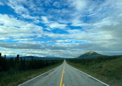 Road aimed straight ahead into the horizon, green grass and trees on both sides and cloudy blue sky above.