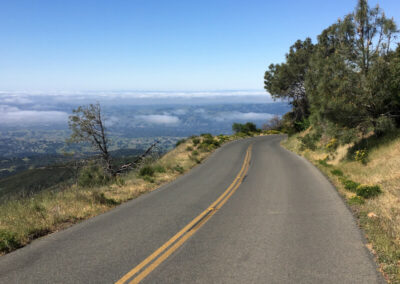 Road winding around mountain, disappearing to the right. Trees on the right and overlooking a valley on the left. Blue sky with some low laying clouds.