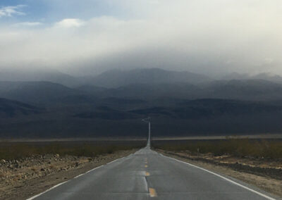 Road aimed straight ahead through the desert and disappearing into dark desert mountains, thick rain clouds above.