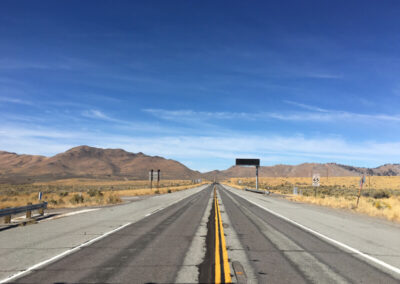 Highway aimed straight ahead into range of desert mountains through desert terrain, blue sky above with some clouds.