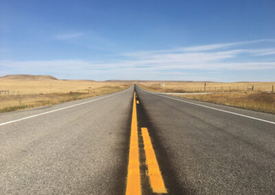 Highway with yellow divider stripes aimed straight ahead through flat yellow grass plains, bright blue sky above with some thin clouds.