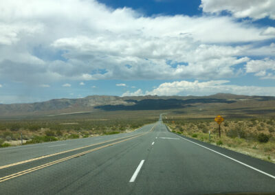 Highway aimed straight ahead into range of desert mountains through desert terrain, blue sky above with many white clouds.