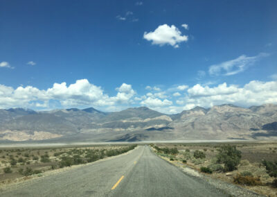 Highway aimed straight ahead into range of desert mountains through desert terrain, blue sky above with some clouds.