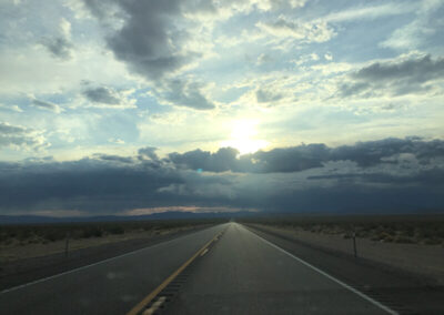 Highway aimed straight ahead into the horizon through desert terrain, cloudy sky with storm clouds far off in distance and sun peering through.