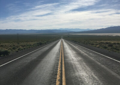 Highway aimed straight ahead into range of desert mountains through desert terrain, blue sky above with some clouds.