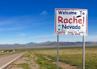 Highway aimed straight ahead into range of desert mountains with green covered desert terrain on both sides, sign at right saying "Welcome to Rachel Nevada. Humans: Yes. Aliens:?" Blue sky above.