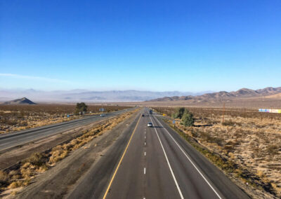 Highway aimed straight ahead into range of desert mountains through desert terrain, blue sky above with some clouds.