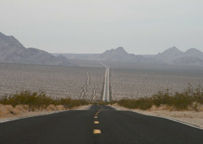 Highway aimed straight ahead into range of desert mountains through barren desert terrain, hazy grey sky above.