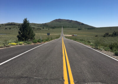 Highway aimed straight ahead into grass covered hills, blue sky above. Some scattered pine trees.