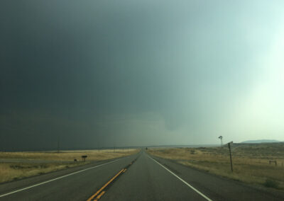 Highway aimed straight ahead through flat grass plains into dark storm clouds, windmill in distance to the right.