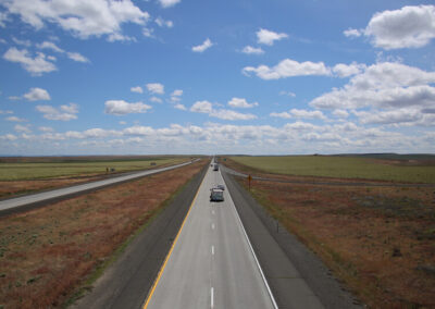 Highway aimed straight ahead over flat Nebraska plains, grazing grass on both sides, blue sky above with some clouds.