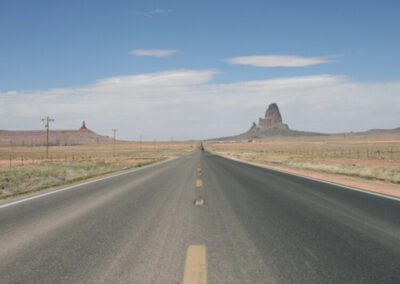 Highway aimed straight ahead into range of desert mountains through desert terrain, with a couple tall rock monuments ahead on both sides of road, blue sky above with some clouds.