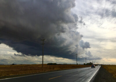 Road aimed straight ahead into the horizon, shiny from recent rain, thick storm clouds above.